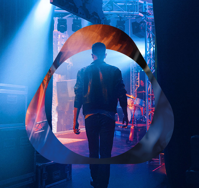 Человек в кожаной куртке за кулисами под синими сценическими прожекторами, наложен логотип.